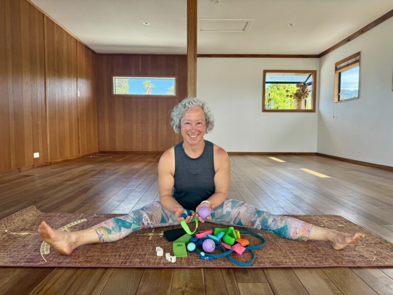 A smiling movement coach sits on a yoga mat in a wide straddle position inside a bright studio, holding colorful foot-mobility tools and props spread out in front of her.