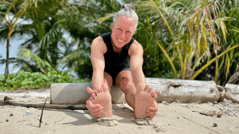 Woman stretching on sandy beach, smiling, holding her sandy feet with palm trees behind her.