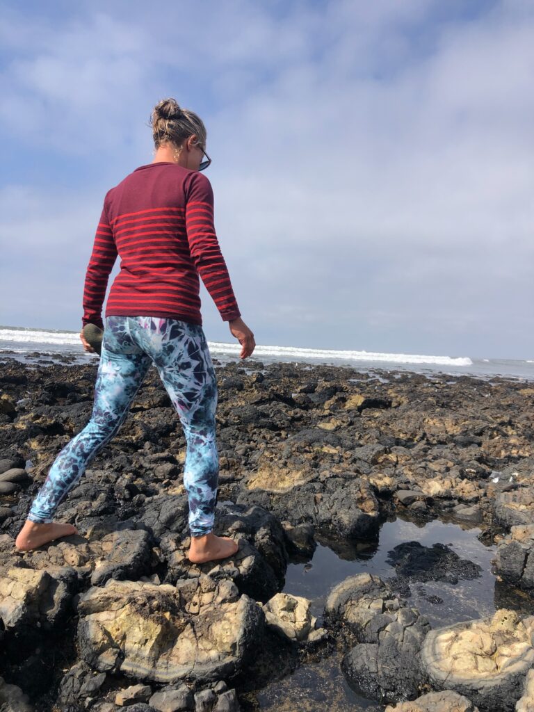 Woman walking barefoot on rocky tide pools, wearing a red striped sweater and patterned leggings.