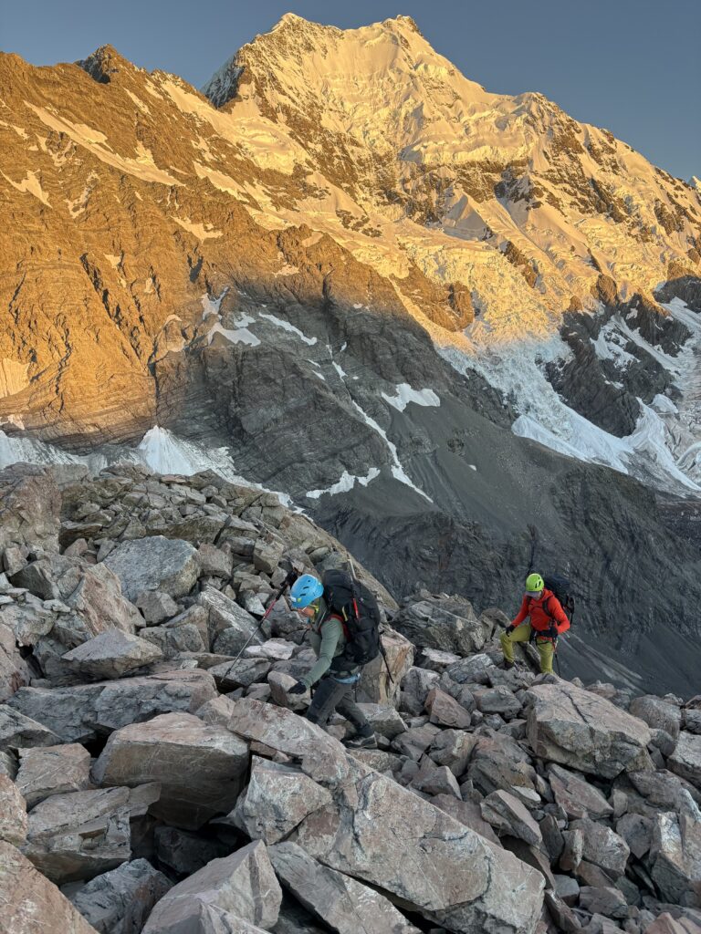 Two mountaineers wearing helmets and backpacks climb over a rocky ridge at sunrise, with a dramatic snow-covered mountain glowing in golden light behind them.