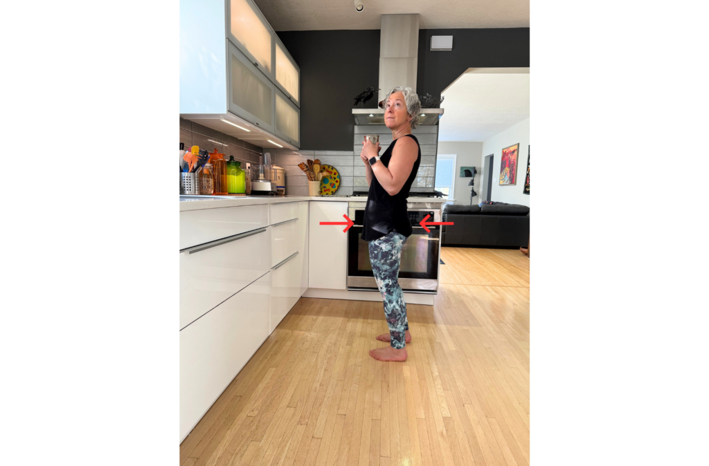 Woman standing barefoot in a modern kitchen holding a mug, shown from the side with red arrows pointing toward her pelvis to illustrate hip alignment.